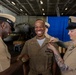 Three U.S. Navy personnel in khaki service uniforms are participating in what appears to be a pinning ceremony inside a ship's hangar bay. The central figure is smiling broadly while two colleagues — one on each side — pin insignia or devices onto his collar or shoulders. A military aircraft and blue ceremonial backdrop are visible in the background.