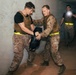 Three individuals in military-style uniforms and tactical vests are inside a dimly lit concrete structure. Two individuals in the foreground appear to be engaged in a close-quarters combat training drill, with one holding a black training pad. A third individual stands in the background illuminating the scene with a handheld flashlight or weapon-mounted light.