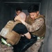 Two individuals in military camouflage uniforms and tactical plate carriers are engaged in what appears to be a close-quarters combat training drill inside a confined corridor or room. A third person is visible in the background observing. The presence of neon green safety markers and a padded striking shield suggests this is a controlled training exercise.