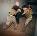 Three individuals in military-style uniforms and tactical vests are engaged in what appears to be a close-quarters combat training exercise inside a concrete corridor or building structure. Two participants are actively grappling with a padded training shield, while a third observes from the background. The setting shows a debris-strewn floor and bare concrete walls consistent with a training facility or mock urban environment.