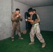 Two military personnel in desert camouflage uniforms and tactical vests are engaged in close-quarters combat training inside a concrete room with artificial grass flooring. One individual holds a padded shield while the other is in a fighting stance. A third person observes in the background.