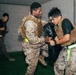 Three individuals in military camouflage uniforms and tactical vests are engaged in what appears to be a close-quarters combat or martial arts training exercise indoors. Two individuals in the foreground are grappling with a black training pad, while a third person is visible in the background. The setting appears to be an indoor training facility with concrete walls and green flooring.
