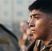 A close-up profile shot of a young male in military camouflage uniform, appearing focused and intense. His face shows visible perspiration, suggesting recent physical exertion. Several other individuals are visible in the blurred background, indicating a group military setting.