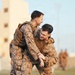 Two military personnel in desert digital camouflage uniforms and tactical vests are engaged in what appears to be a hand-to-hand combat training drill on a grass field. Both individuals are smiling, indicating a training or practice context rather than a hostile encounter. Additional personnel in similar uniforms are visible in the blurred background near a tan-colored building.