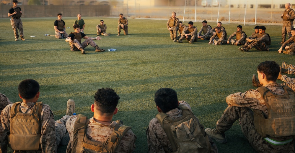 A large group of military personnel in camouflage uniforms are gathered on a green grass field, arranged in a semicircle or circle formation. One individual appears to be addressing or instructing the group from a central position on the ground. The setting appears to be a military base or training facility, photographed during golden hour lighting conditions.