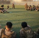 A large group of military personnel in camouflage uniforms are gathered on a green grass field, arranged in a semicircle or circle formation. One individual appears to be addressing or instructing the group from a central position on the ground. The setting appears to be a military base or training facility, photographed during golden hour lighting conditions.