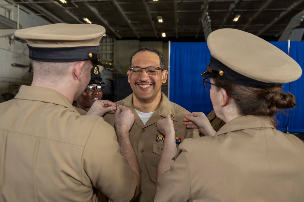 A U.S. Navy promotion ceremony is taking place in what appears to be a ship's hangar bay. Two sailors are simultaneously pinning new rank insignia onto the collar of a smiling servicemember, a traditional 'pinning' ritual marking an advancement in rank. A fourth sailor observes the ceremony in the background near a blue curtain backdrop.