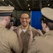 A U.S. Navy promotion ceremony is taking place in what appears to be a ship's hangar bay. Two sailors are simultaneously pinning new rank insignia onto the collar of a smiling servicemember, a traditional 'pinning' ritual marking an advancement in rank. A fourth sailor observes the ceremony in the background near a blue curtain backdrop.