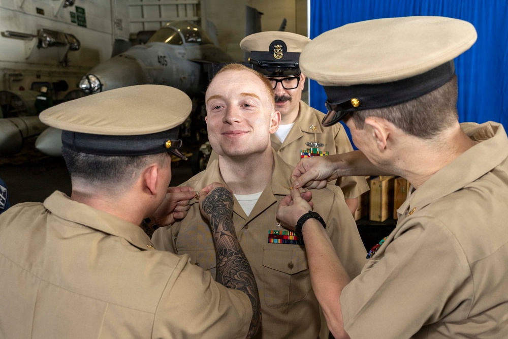 A U.S. Navy promotion or pinning ceremony is taking place in what appears to be an aircraft hangar bay aboard a naval vessel. A young sailor with red hair and a sleeve tattoo is being pinned by two fellow sailors while a senior petty officer observes from behind. Military aircraft are visible in the background.