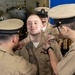 A U.S. Navy promotion or pinning ceremony is taking place in what appears to be an aircraft hangar bay aboard a naval vessel. A young sailor with red hair and a sleeve tattoo is being pinned by two fellow sailors while a senior petty officer observes from behind. Military aircraft are visible in the background.
