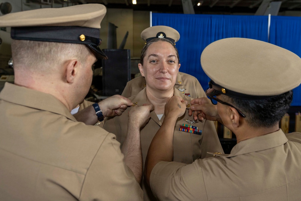 A U.S. Navy promotion or pinning ceremony is taking place, with two service members simultaneously pinning new insignia onto the collar or chest area of a female sailor. The recipient is wearing a khaki uniform with multiple service ribbons and qualification badges, and all three individuals are in U.S. Navy Service Uniform (NSU) khaki attire. The ceremony appears to be conducted aboard a naval vessel or in a military facility with a blue curtain backdrop.