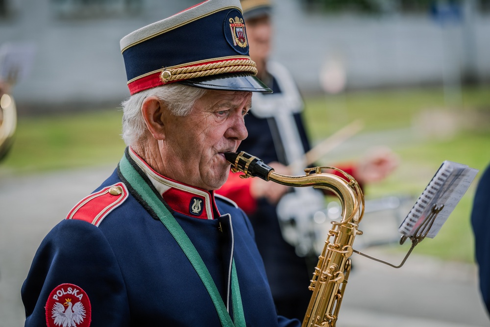 An elderly man in a Polish military-style band uniform is playing a tenor saxophone outdoors. He is reading from sheet music on a stand while performing, likely as part of a marching or ceremonial band. Additional band members and instruments are visible in the blurred background.
