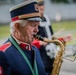 An elderly man in a Polish military-style band uniform is playing a tenor saxophone outdoors. He is reading from sheet music on a stand while performing, likely as part of a marching or ceremonial band. Additional band members and instruments are visible in the blurred background.