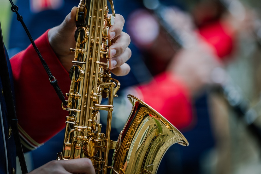 A close-up photograph of a person playing a gold-lacquered saxophone, likely during a band performance or parade. The player is wearing a red uniform jacket, suggesting membership in a marching band or ensemble. Additional blurred figures in similar attire are visible in the background.