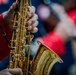 A close-up photograph of a person playing a gold-lacquered saxophone, likely during a band performance or parade. The player is wearing a red uniform jacket, suggesting membership in a marching band or ensemble. Additional blurred figures in similar attire are visible in the background.