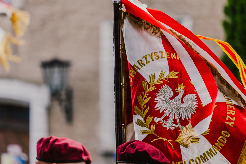 The image shows a ceremonial banner or standard featuring the Polish White Eagle emblem on a red and white background, consistent with Polish national colors. Partial text visible includes 'STOWARZYSZENIE' (Association) and 'COMMANDO' along with 'ŻOŁNIERZY' (Soldiers), suggesting a Polish veterans or military association standard. At least one person wearing a dark burgundy/maroon beret is partially visible in the foreground.