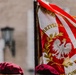 The image shows a ceremonial banner or standard featuring the Polish White Eagle emblem on a red and white background, consistent with Polish national colors. Partial text visible includes 'STOWARZYSZENIE' (Association) and 'COMMANDO' along with 'ŻOŁNIERZY' (Soldiers), suggesting a Polish veterans or military association standard. At least one person wearing a dark burgundy/maroon beret is partially visible in the foreground.