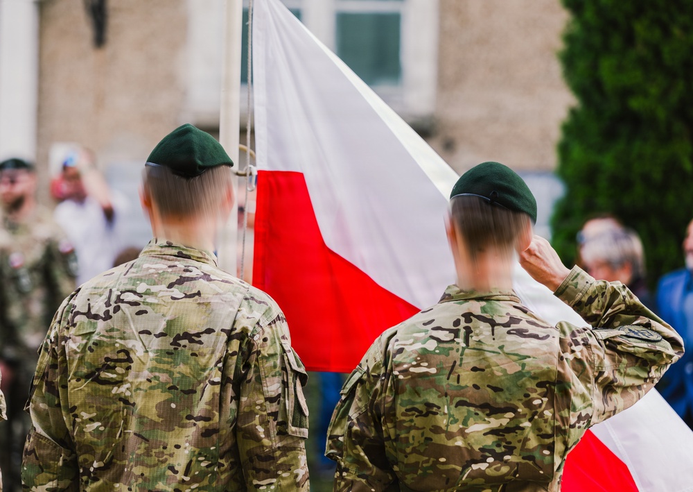 Two soldiers in camouflage uniforms and dark green berets stand at attention before a Polish national flag (white and red horizontal stripes). One soldier is visibly saluting. The scene appears to be a formal military ceremony taking place outdoors in front of a building.