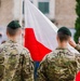 Two soldiers in camouflage uniforms and dark green berets stand at attention before a Polish national flag (white and red horizontal stripes). One soldier is visibly saluting. The scene appears to be a formal military ceremony taking place outdoors in front of a building.