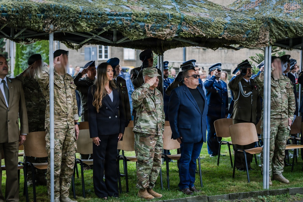 A formal military ceremony is taking place outdoors under a camouflage netting canopy. Multiple military personnel in various uniforms are standing at salute alongside civilians in formal attire. The setting appears to be an official commemorative or ceremonial event with mixed military and civilian attendees.