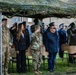 A formal military ceremony is taking place outdoors under a camouflage netting canopy. Multiple military personnel in various uniforms are standing at salute alongside civilians in formal attire. The setting appears to be an official commemorative or ceremonial event with mixed military and civilian attendees.