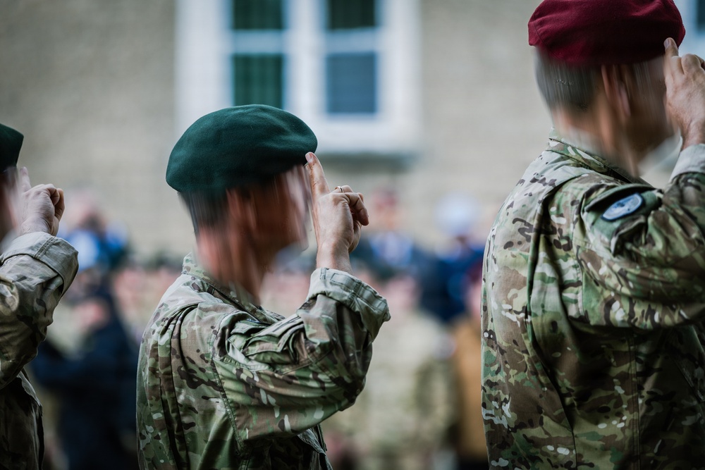 Military personnel in camouflage uniforms are performing a salute during what appears to be a formal parade or ceremony. At least two distinct beret colors are visible — green and maroon — suggesting personnel from different units or branches. A crowd of observers is visible in the blurred background near a stone building.