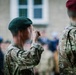Military personnel in camouflage uniforms are performing a salute during what appears to be a formal parade or ceremony. At least two distinct beret colors are visible — green and maroon — suggesting personnel from different units or branches. A crowd of observers is visible in the blurred background near a stone building.