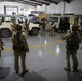 A group of heavily armed and equipped military personnel are gathered inside a large indoor facility, appearing to receive a briefing or conduct a pre-mission coordination. The soldiers are wearing desert camouflage uniforms and full tactical gear including helmets, body armor, and weapons. Several military and civilian-style vehicles are parked in the background of what appears to be a motor pool or staging area.