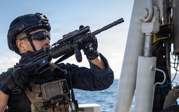 A heavily equipped tactical operator is positioned on the deck of a naval or coast guard vessel, aiming a rifle toward an unseen target. The individual is wearing full tactical gear including a plate carrier, helmet with night vision mount, and protective eyewear. The open ocean is visible in the background, suggesting an at-sea security or boarding operation.
