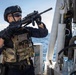 A heavily equipped tactical operator is positioned on the deck of a naval or coast guard vessel, aiming a rifle toward an unseen target. The individual is wearing full tactical gear including a plate carrier, helmet with night vision mount, and protective eyewear. The open ocean is visible in the background, suggesting an at-sea security or boarding operation.
