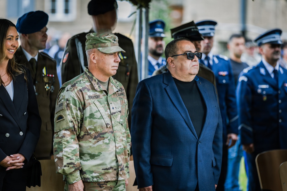A formal military ceremony featuring personnel from multiple branches and nations. A U.S. Army officer with the name tape 'DEMONTIGNY' and two-star general rank insignia stands prominently in the foreground alongside a civilian in a blue blazer. Behind them are rows of uniformed personnel, including what appear to be Romanian Air Force officers in blue dress uniforms.