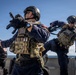 Three armed tactical personnel are conducting what appears to be a maritime boarding or vessel interdiction drill on the deck of a ship. The operators are wearing tactical gear including plate carriers, helmets, and are carrying firearms. One operator is climbing over a ship railing while the others provide cover, suggesting a coordinated boarding exercise.