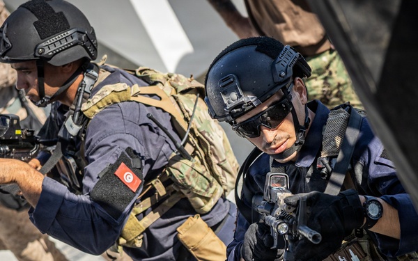 The image shows armed military or special operations personnel conducting what appears to be a tactical training exercise or boarding operation. At least two operators in the foreground are wearing tactical gear, helmets, and carrying firearms in a ready position. A Tunisian flag patch is visible on one operator's sleeve, suggesting multinational military cooperation or joint training.