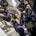 The image shows armed military or special operations personnel conducting what appears to be a tactical training exercise or boarding operation. At least two operators in the foreground are wearing tactical gear, helmets, and carrying firearms in a ready position. A Tunisian flag patch is visible on one operator's sleeve, suggesting multinational military cooperation or joint training.
