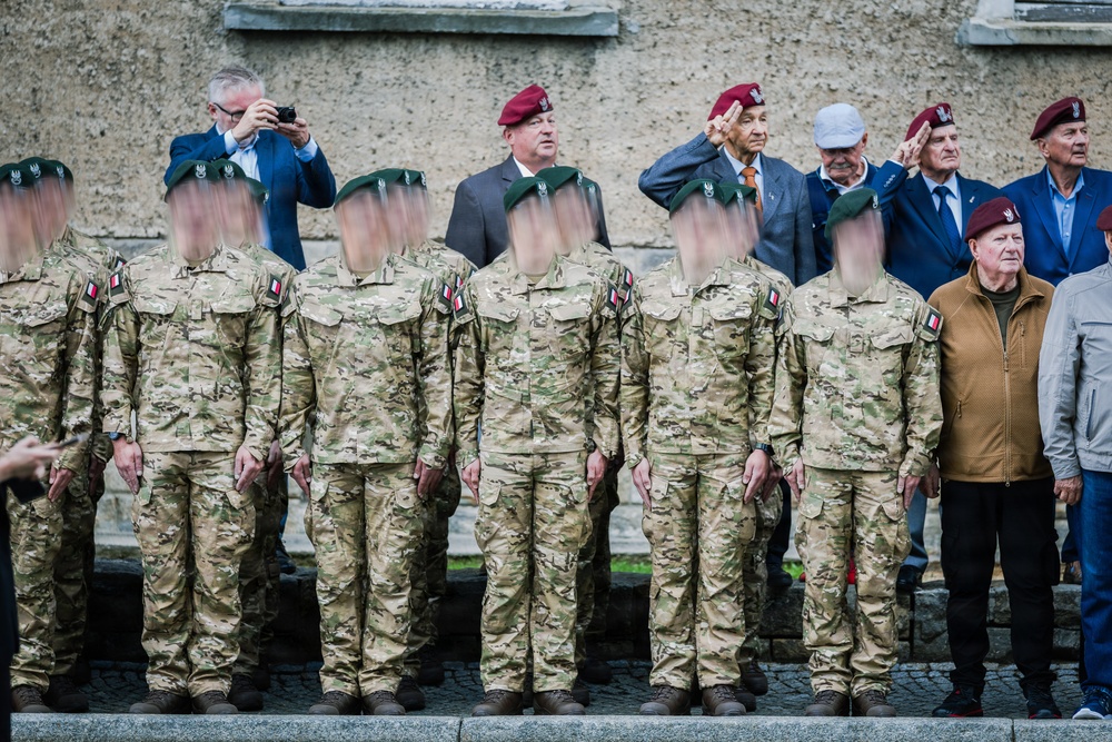 A formal military ceremony featuring a row of soldiers in multicam camouflage uniforms wearing green berets, standing at attention. Behind them are older individuals, some wearing maroon/burgundy berets, likely veterans, with several saluting. A civilian in a blue suit is photographing the event.