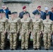A formal military ceremony featuring a row of soldiers in multicam camouflage uniforms wearing green berets, standing at attention. Behind them are older individuals, some wearing maroon/burgundy berets, likely veterans, with several saluting. A civilian in a blue suit is photographing the event.