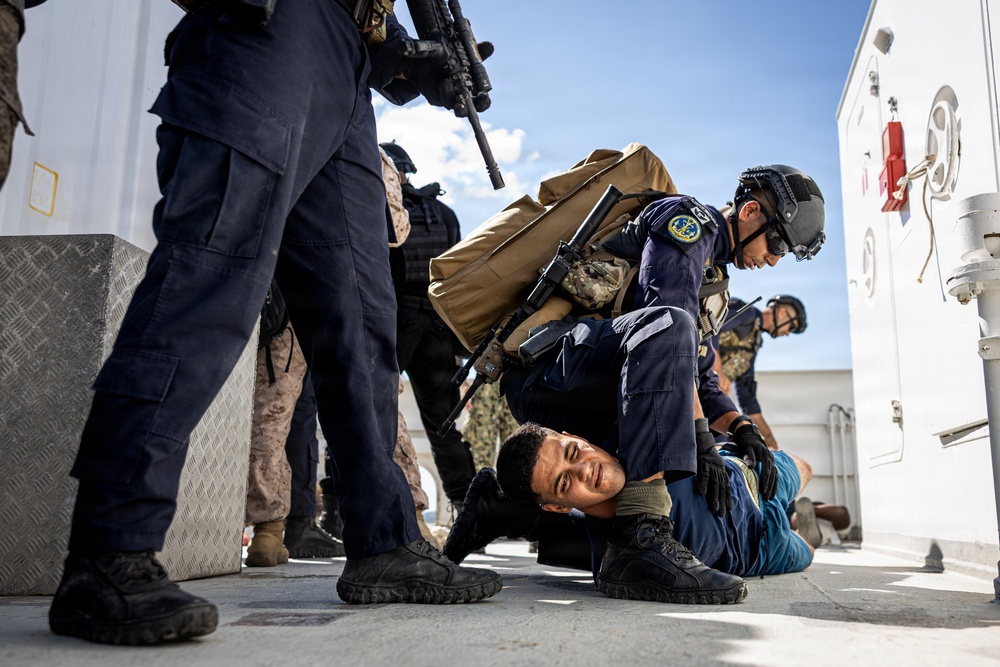The image depicts what appears to be a military or law enforcement training exercise on the deck of a naval vessel. Armed personnel in dark blue uniforms and tactical gear are restraining a person on the ground, while others stand nearby with weapons. The scene includes multiple personnel with different uniforms suggesting a joint or multinational exercise.