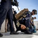 The image depicts what appears to be a military or law enforcement training exercise on the deck of a naval vessel. Armed personnel in dark blue uniforms and tactical gear are restraining a person on the ground, while others stand nearby with weapons. The scene includes multiple personnel with different uniforms suggesting a joint or multinational exercise.