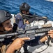 Two armed military or law enforcement personnel are positioned on the deck of a naval vessel, holding assault rifles in a ready stance. They are wearing tactical gear including ballistic helmets, body armor, and gloves, suggesting a maritime security or boarding operation. The setting is an open sea environment with a coastline visible in the background.