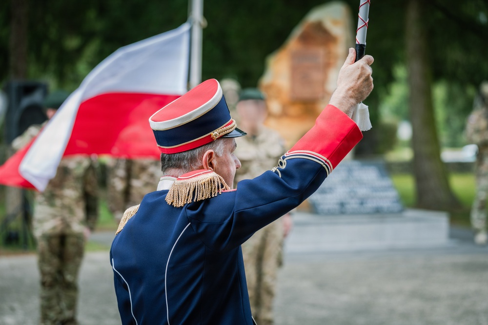 A military officer in formal dress uniform is conducting or leading a ceremony, raising a baton or staff with one arm. A red-and-white flag, consistent with the Polish national flag, is visible in the background. Several soldiers in camouflage uniforms and a stone monument are visible in the blurred background.