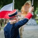 A military officer in formal dress uniform is conducting or leading a ceremony, raising a baton or staff with one arm. A red-and-white flag, consistent with the Polish national flag, is visible in the background. Several soldiers in camouflage uniforms and a stone monument are visible in the blurred background.