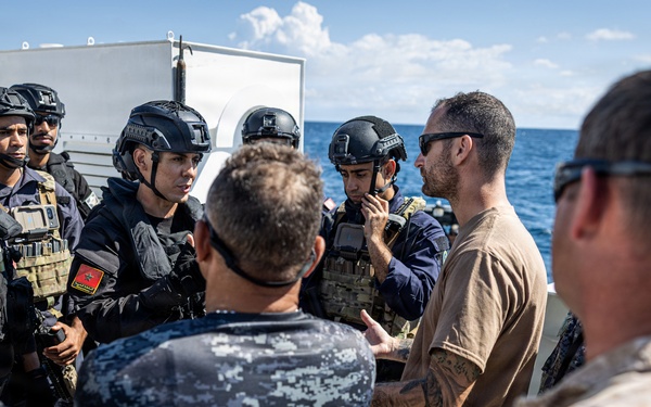 A group of military and security personnel are gathered on the deck of a naval vessel at sea, engaged in what appears to be a briefing or coordination meeting. The group includes personnel in different uniforms, suggesting a multinational or joint operation context. Some personnel are wearing tactical gear including helmets, body armor, and visible weapons.