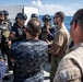 A group of military and security personnel are gathered on the deck of a naval vessel at sea, engaged in what appears to be a briefing or coordination meeting. The group includes personnel in different uniforms, suggesting a multinational or joint operation context. Some personnel are wearing tactical gear including helmets, body armor, and visible weapons.