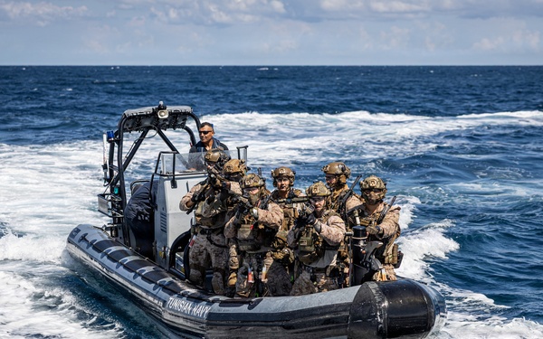 A rigid inflatable boat (RIB) marked 'TUNISIAN NAVY' carries a group of armed military personnel in full tactical gear across open ocean waters. The soldiers are equipped with helmets, body armor, and weapons, suggesting a maritime special operations or boarding exercise. The boat is moving at speed, creating a visible wake behind it.