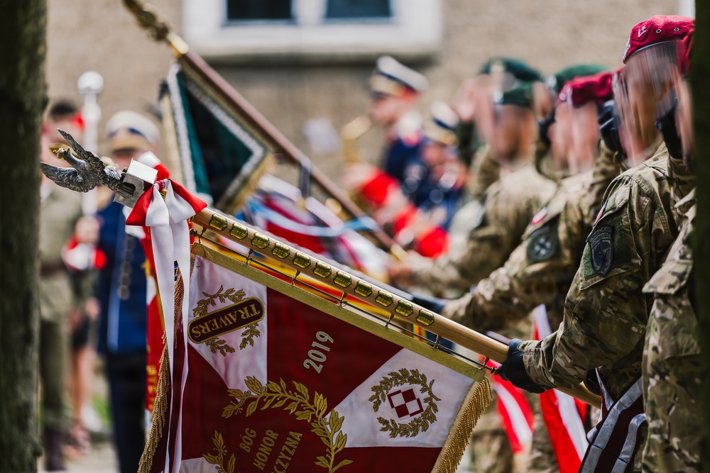 A military ceremony featuring uniformed personnel holding ceremonial standards and flags. The prominent banner in the foreground displays the text 'TRAWERS', '2019', 'BÓG HONOR OJCZYZNA' (God, Honor, Fatherland) along with Polish military insignia including a cross and laurel wreath. The scene includes a military band and multiple service members in various uniforms in the background.