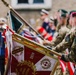 A military ceremony featuring uniformed personnel holding ceremonial standards and flags. The prominent banner in the foreground displays the text 'TRAWERS', '2019', 'BÓG HONOR OJCZYZNA' (God, Honor, Fatherland) along with Polish military insignia including a cross and laurel wreath. The scene includes a military band and multiple service members in various uniforms in the background.