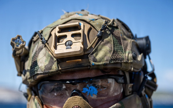 A heavily equipped military or tactical operator is photographed in close-up against a blue water and sky background. The individual is wearing full tactical gear including a multicam helmet with multiple attachments, protective eyewear with visible blue paint/dye splatter, and a mesh lower face mask. The blue paint on the goggles suggests participation in a training exercise using simulated munitions.