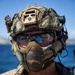 A heavily equipped military or tactical operator is photographed in close-up against a blue water and sky background. The individual is wearing full tactical gear including a multicam helmet with multiple attachments, protective eyewear with visible blue paint/dye splatter, and a mesh lower face mask. The blue paint on the goggles suggests participation in a training exercise using simulated munitions.