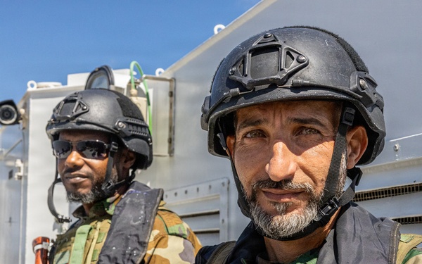 Two armed military personnel are positioned on what appears to be the deck of a vessel at sea. Both wear camouflage uniforms, tactical helmets, and body armor, with visible Libyan national flag patches and a military unit insignia. The foreground figure also wears an 'MST' branded life vest, suggesting a maritime security or naval context.