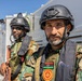 Two armed military personnel are positioned on what appears to be the deck of a vessel at sea. Both wear camouflage uniforms, tactical helmets, and body armor, with visible Libyan national flag patches and a military unit insignia. The foreground figure also wears an 'MST' branded life vest, suggesting a maritime security or naval context.